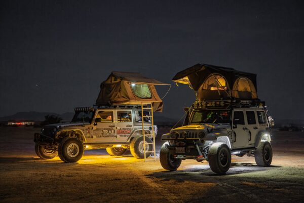 Two overland-built Jeeps are parked in the desert with lights illuminating their undercarriages.