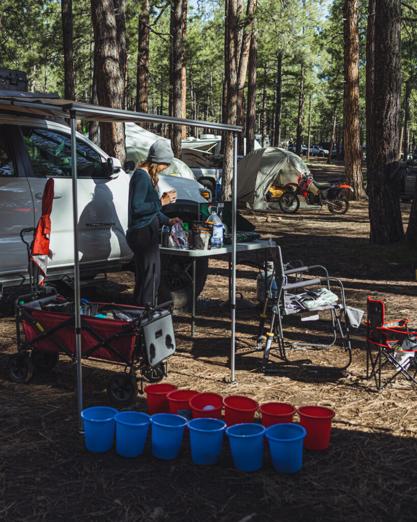 Overland Expo camper preparing for the day by making breakfast while drinking coffee. She is wearing a hat and thermal clothes for the crisp morning, but the bright sun tells us it will likely warm up.